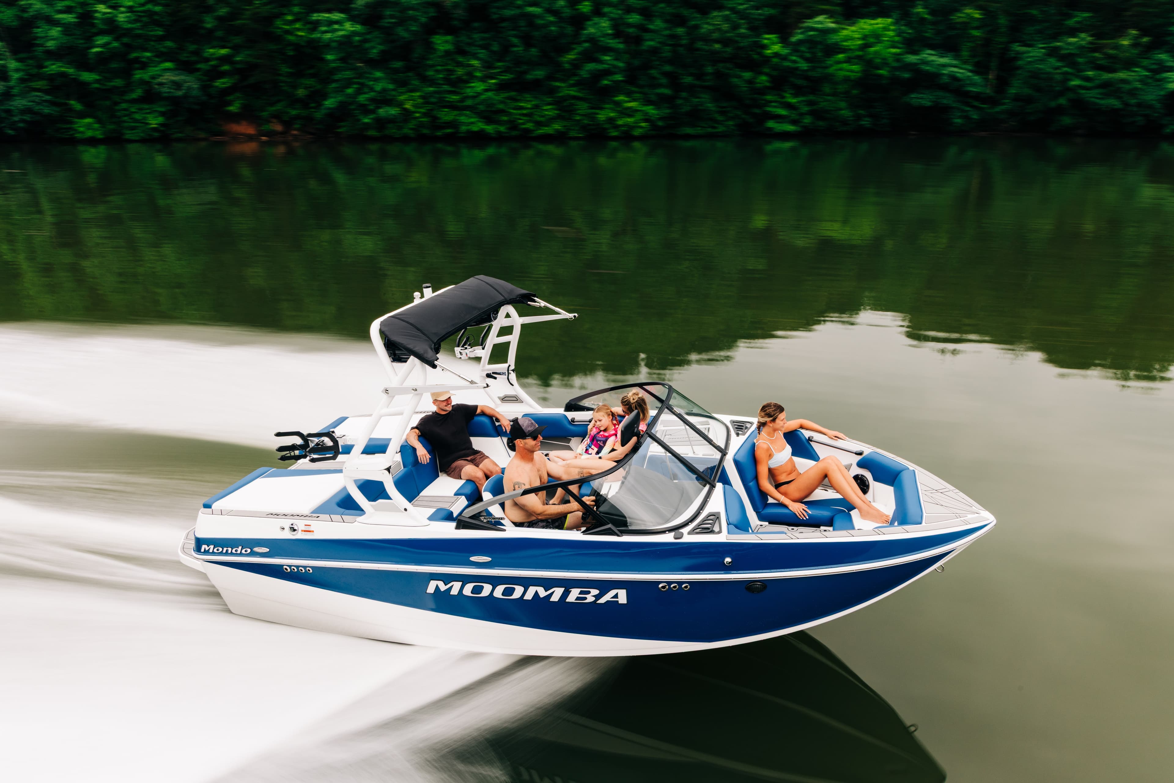 A Moomba wakeboard boat with blue and white hull underway on water carrying six passengers, with a wakeboard tower and forested shoreline in the background.